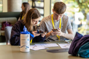 Two young people work together at a desk, taking photos of a piece of paper with their phones.