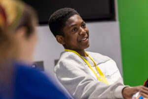 A young person wearing a yellow lanyard sits in a classroom, smiling.