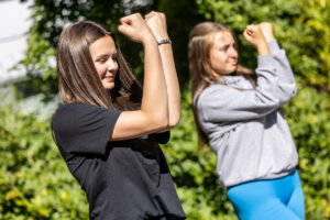 Two young people perform a choreographed dance outside with their arms flexed in a power pose in front of their faces. The background is full of bright greenery.