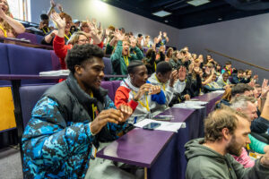 An auditorium filled with young people following along in a group game.