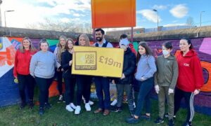 A group of young people stand outside in front of a brick wall with a colourful mural. They hold a large yellow sign that reads '£19m' with the CashBack for Communities logo.
