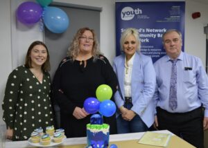 The Awards Team stands smiling in front of a table with cupcakes, balloons and a blue tiered cake decorated with '20 DYA'