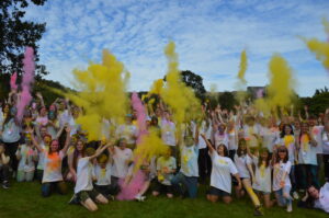 A large group of young people wearing white t-shirts pose in a grassy field. The group throw large clouds of yellow and pink colour powder into the air.