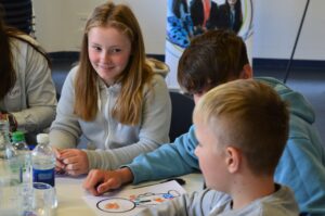 Three young people are seated at a table colouring on worksheets.