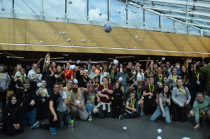 A crowd of young people are gathered in a glass-ceiling auditorium. They are throwing balls of paper towards the camera.