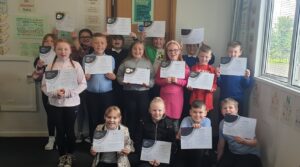 A group of young people stand in a classroom, holding up Youth Scotland Award certificates and smiling for the camera.