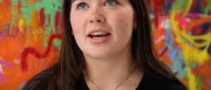 Abbie, a member of KYTHE sits before a colourful painted wall