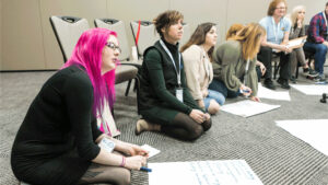 A group of youth workers sit on the grey carpet of a conference room, looking off camera. A row of chairs is lined up behind them and on the floor in front of them are large white sheets of paper with handwriting.