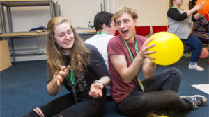 Two young people sit on the floor of a classroom back to back. They are smiling and passing a yellow balloon between them.