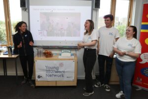 A group of young people in matching white t-shirts stand at the front of a classroom next to a projector screen. On the table in front of them is a banner with the logo for the Young Islanders Network.