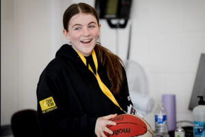 A girl wearing a Reach hoodie holds a basketball while smiling at someone