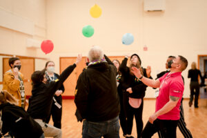 A group of young people and youth workers stand in circle a gymnasium, tossing colourful balloons in the air.