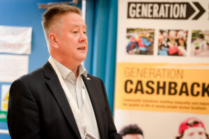Keith Brown stands speaking in front of a pop up banner that reads 'Generation CashBack'