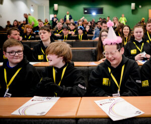 A group of young people sit in rows of desks in an auditorium. They are wearing matching black Reach sweatshirts and yellow lanyards.