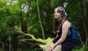 A young person sits on a moss-covered log, looking off into a green forest.