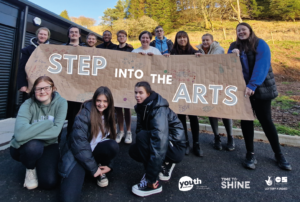 Thirteen young people stand and kneel in a group outside in front of a sunny hillside. They hold a large brown paper banner, which has white text reading 'Step into the Arts' digitally added to it.