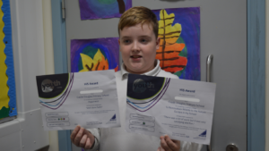 A young person stands in a school classroom holding two certificates for Youth Scotland Hi5 Awards.