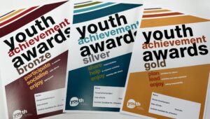 Booklets for the Youth Achievement Awards Bronze, Silver, and Gold levels are arranged in a fan, in brown, blue and gold respectively.