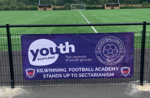 A banner attached to a football stand, with the Youth Scotland and #FOOTBALLUNITES logos reads "Kilwinning Football Academy stands up to sectarianism"