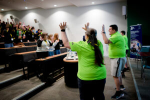Several youth workers in neon green t-shirts stand at the front of an auditorium leading an icebreaker for a group of young people.