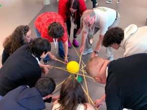 A group of youth workers and young people complete a team-building challenge with bamboo sticks and a yellow play ball.