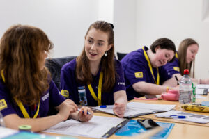 A group of young people wearing purple polo shirts sit at a large conference table working on Dynamic Youth Award booklets.