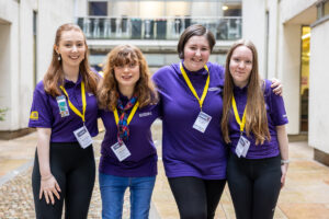 Four young people wearing purple polo shirts and yellow lanyards smile for the camera.