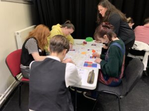 A youth worker trainer supports youth workers in their learning around a desk full of colourful crafts materials