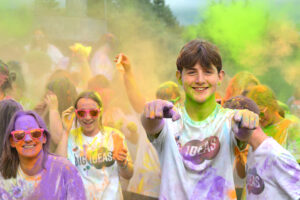 A young person points at the camera as coloured dust is thrown in the air all around them.